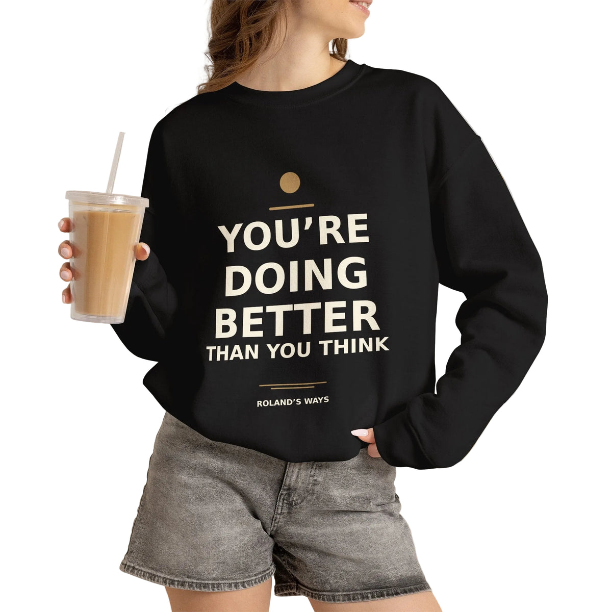 Model holding an iced drink while wearing the black You’re Doing Better Than You Think crewneck with denim shorts.