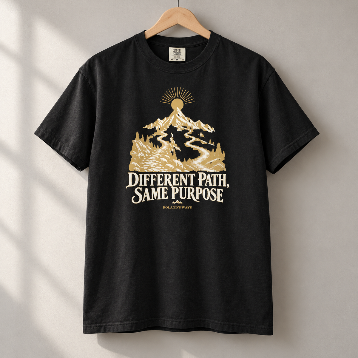 AI mockup of the hanging black Different Path, Same Purpose T-shirt with gold and ivory mountain artwork.