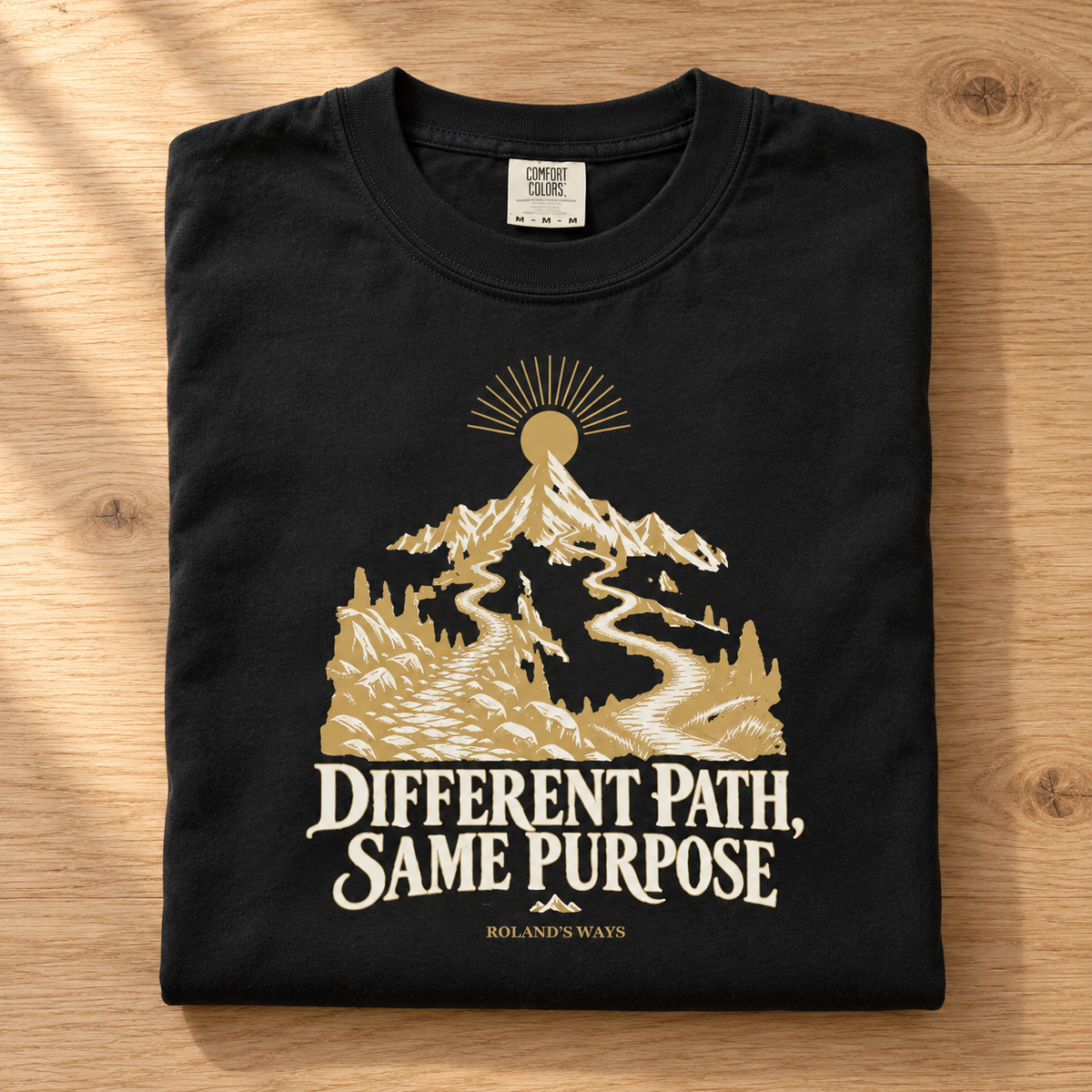 AI mockup of a folded shirt on pale wood: the black Different Path, Same Purpose T-shirt with gold and ivory mountain artwork.