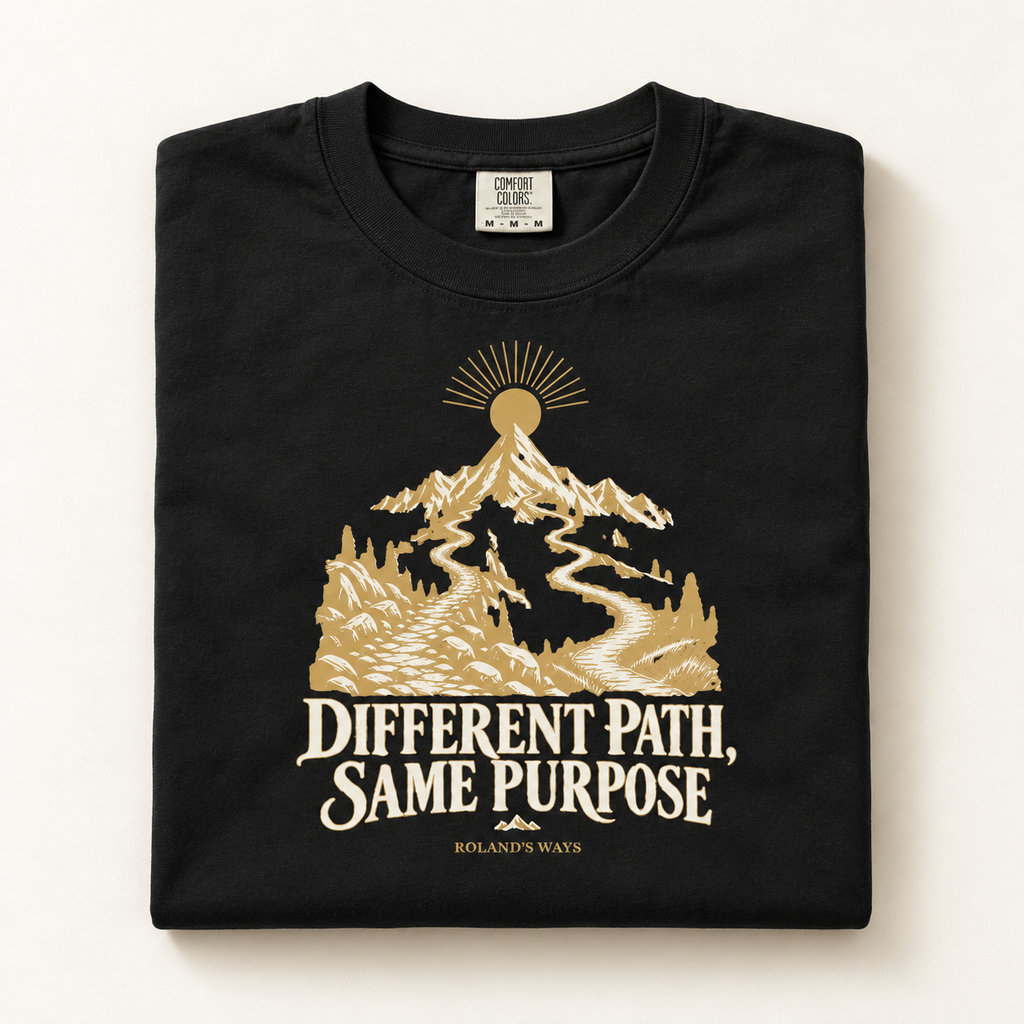 AI mockup of a folded shirt on an ivory surface: the black Different Path, Same Purpose T-shirt with gold and ivory mountain artwork.