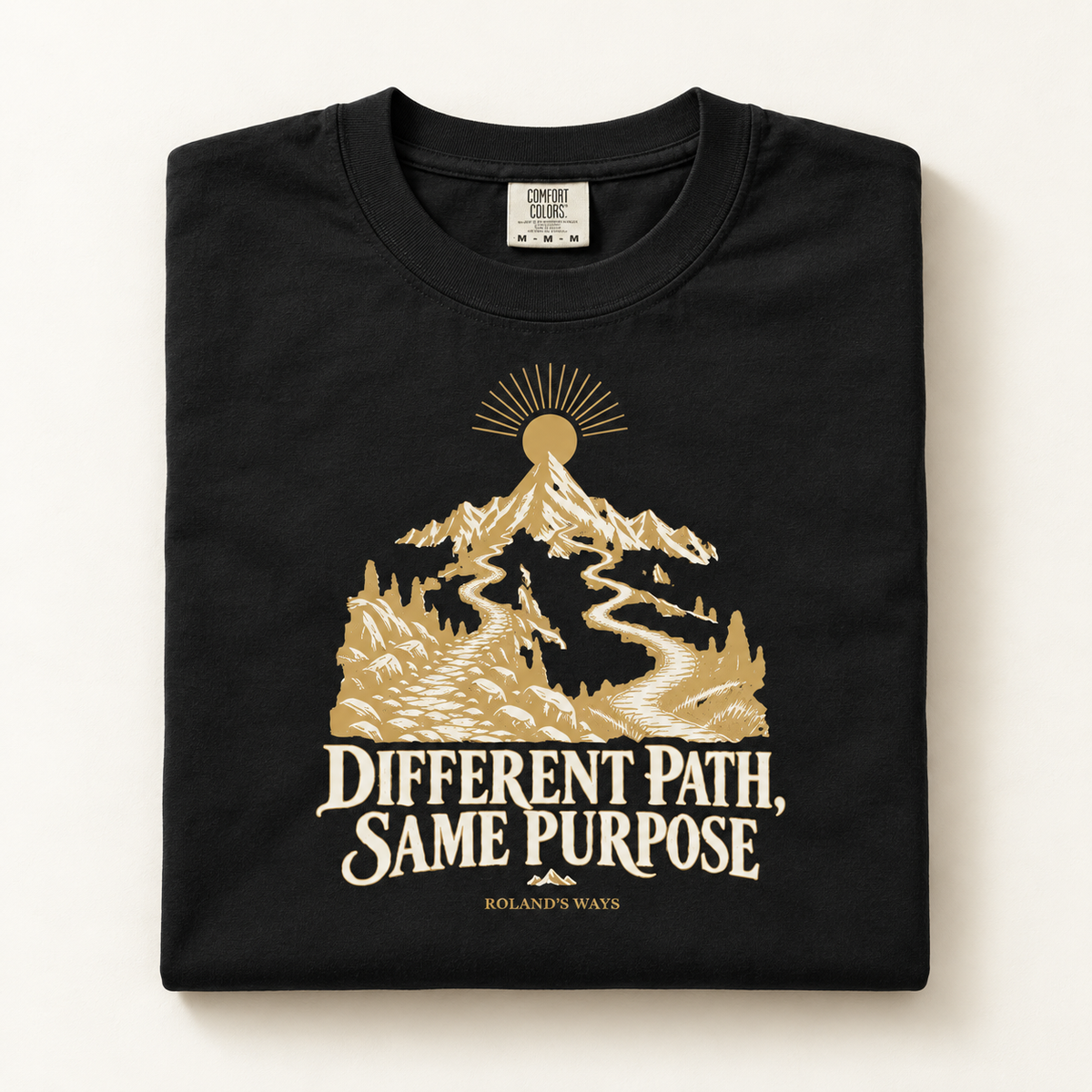 AI mockup of a folded shirt on an ivory surface: the black Different Path, Same Purpose T-shirt with gold and ivory mountain artwork.